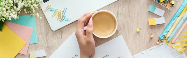 Vista cortada de mulher segurando copo com café na mesa de madeira com laptop e artigos de papelaria — Fotografia de Stock