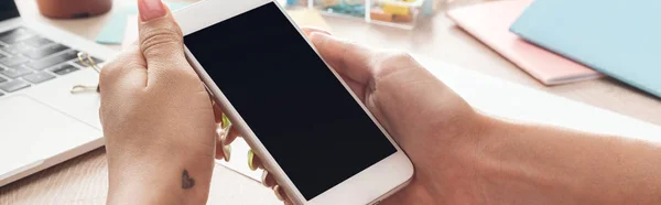 Vista recortada de la mujer sosteniendo el teléfono inteligente en las manos sobre la mesa de madera con ordenador portátil y papelería - foto de stock