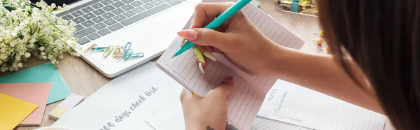 Ausgeschnittene Ansicht einer Frau, die Notizen in Notizblock schreibt, hinter einem Holztisch mit Laptop, Planern und Schreibwaren sitzt — Stockfoto