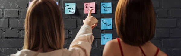 Vista posterior de las mujeres que señalan con el dedo en almohadillas adhesivas de colores con notas en la pared de ladrillo negro - foto de stock