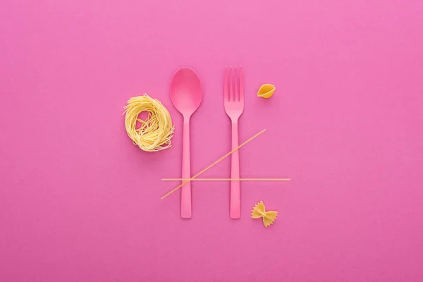 Рожева пластикова ложка і виделка біля різних видів макаронних виробів ізольовані на рожевому — стокове фото