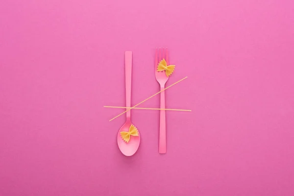 Pink plastic spoon upside down and fork, spaghetti and two uncooked macaroni isolated on pink — Stock Photo