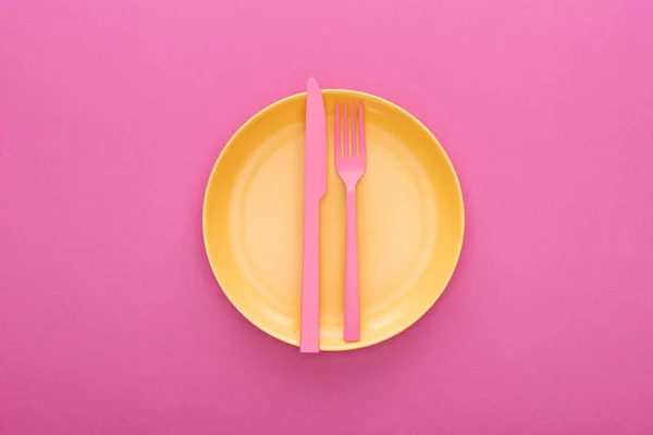 Вид сверху на розовую пластиковую вилку и нож на желтую пластиковую пластину на розовом фоне — стоковое фото