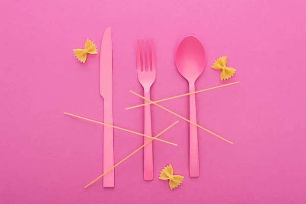 Cuchillo de plástico rosa, tenedor y cuchara, espaguetis y pasta farfalle sin cocer aislados en rosa - foto de stock