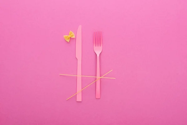 Coltello e forchetta in plastica rosa, spaghetti e farfalle crude isolate su rosa — Foto stock