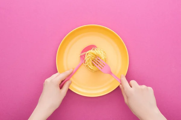 Ausgeschnittene Ansicht eines Erwachsenen, der einen rosa Löffel hält und Vermicelli-Pasta mit Gabel auf gelbem Plastikteller auf rosa Hintergrund nimmt — Stockfoto