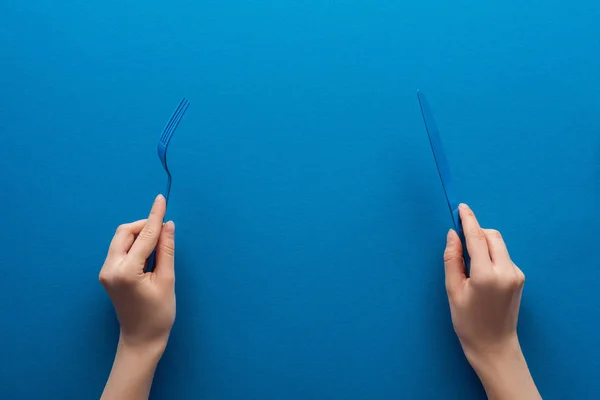 Vue recadrée de la femme tenant fourchette et couteau en plastique bleu sur fond bleu — Photo de stock
