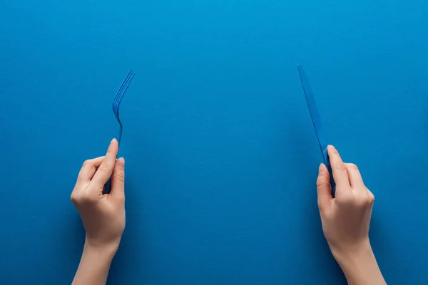 Vista recortada de la mujer sosteniendo tenedor de plástico azul y cuchillo sobre fondo azul - foto de stock