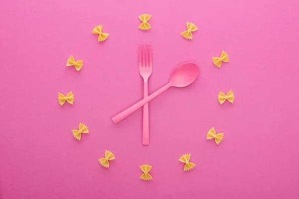 Top view of clock concept with uncooked farfalle pasta, plastic fork and spoon in middle isolated on pink — Stock Photo
