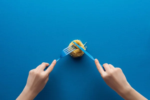 Vista recortada de la mujer sosteniendo tenedor de plástico azul y corte de pasta vermicelli con cuchillo sobre fondo azul - foto de stock
