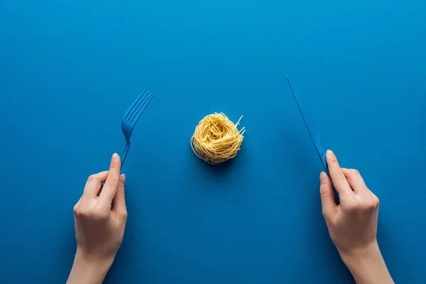 Cropped view of woman holding blue plastic fork and knife with vermicelli pasta in middle on blue background — Stock Photo
