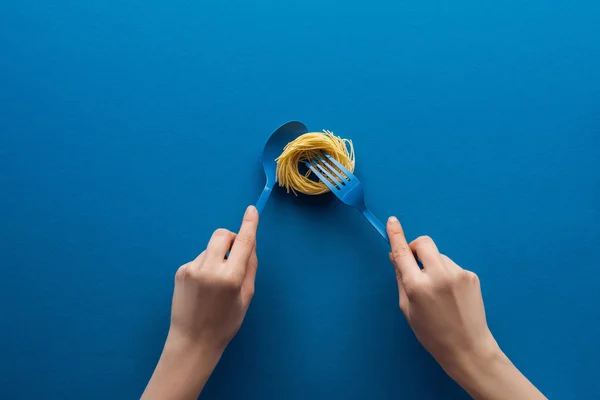 Vista recortada de la mujer sosteniendo cuchara azul y tomando pasta vermicelli con tenedor aislado en azul - foto de stock