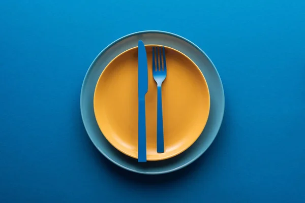 Top view of blue plastic knife and fork on yellow plate above another plate on blue background — Stock Photo