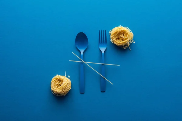 Вид зверху блакитної пластикової ложки і виделки з спагетті біля вермішельської пасти на синьому фоні — стокове фото