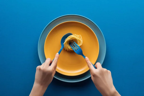 Vista recortada de la mujer sosteniendo cuchara azul y tomando pasta vermicelli con tenedor en plato amarillo por encima de otro plato sobre fondo azul - foto de stock