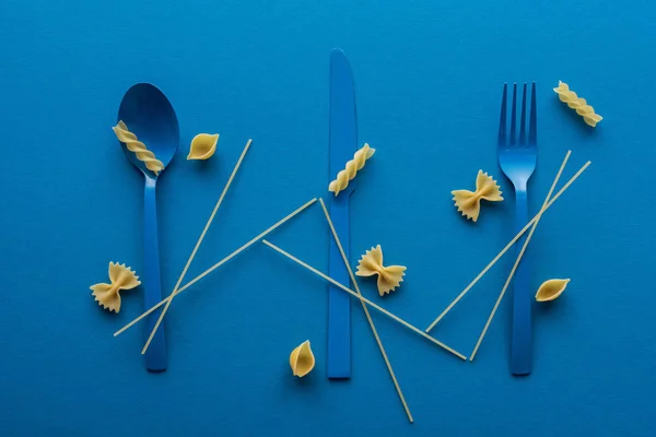 Cubiertos de plástico azul con espaguetis sin cocer y diferentes tipos de pasta sobre fondo azul - foto de stock