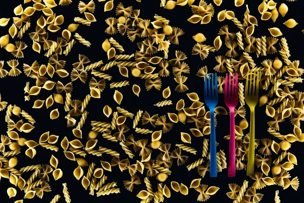 Top view of plastic colorful forks on mixed uncooked pasta isolated on black — Stock Photo