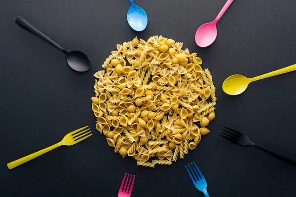 Draufsicht auf ungekochte Pasta umgeben von buntem Besteck auf schwarzem Hintergrund — Stockfoto