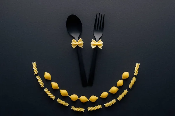 Pâtes farfalle non cuites sur cuillère et fourchette en plastique près des coquilles macaroni et rotini pâtes isolées sur noir — Photo de stock