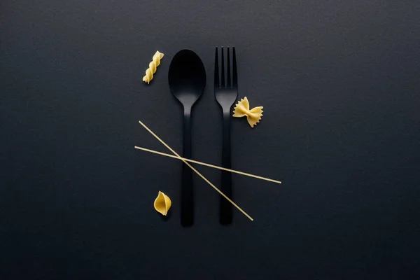 Чорна пластикова ложка, виделка та чотири різні види макаронних виробів на чорному тлі — стокове фото