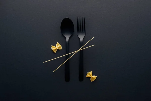 Пластмассовая ложка и вилка с фарфоровой пастой и спагетти на черном фоне — стоковое фото