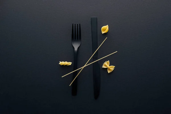 Black plastic knife upside down, fork and four different kinds of pasta on black background — Stock Photo
