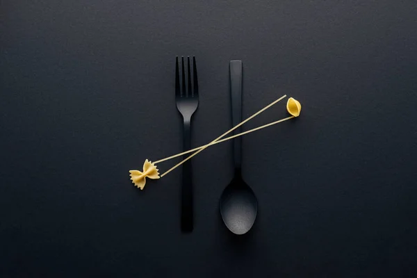 Flat lay of spaghetti, farfalle pasta and shell macaroni on spoon upside down and fork on black background — Stock Photo