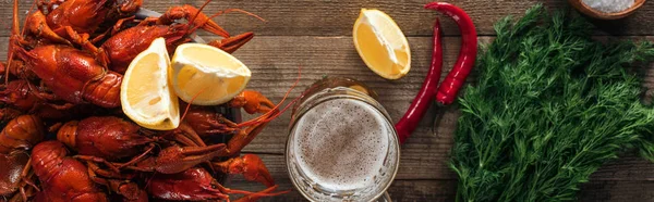 Panoramic shot of red lobsters, dill, pepper, lemon slices and glass with beer on wooden surface — Stock Photo