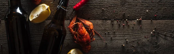 Panoramic shot of red lobsters, lemon slices, peppers and bottles with beer on wooden surface — Stock Photo