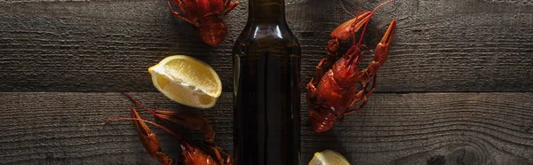 Plano panorámico de langostas rojas, rodajas de limón y botella de vidrio con cerveza en la superficie de madera - foto de stock