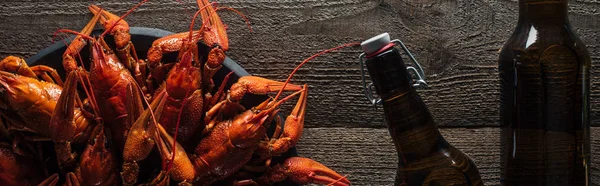 Tiro panorâmico de lagostas vermelhas na placa e garrafas de vidro com cerveja na superfície de madeira — Fotografia de Stock