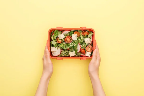 Vista ritagliata di donna che tiene il pranzo scatola con insalata in mano su sfondo giallo — Foto stock