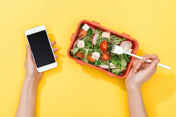Cropped view of woman with fork and lunch box with salad using smartphone — Stock Photo