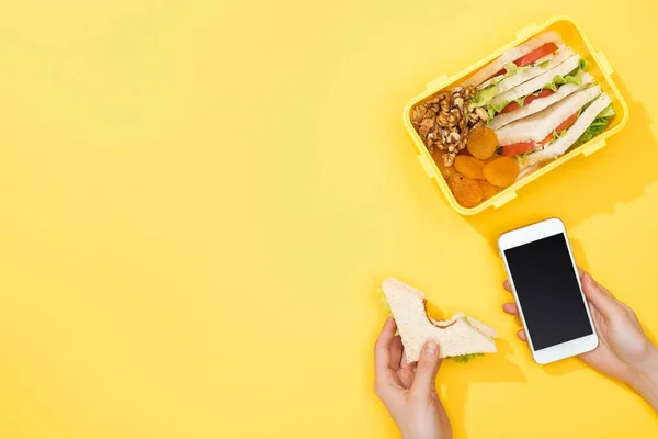 Cropped view of woman holding sandwich and smartphone near lunch box with food — Stock Photo