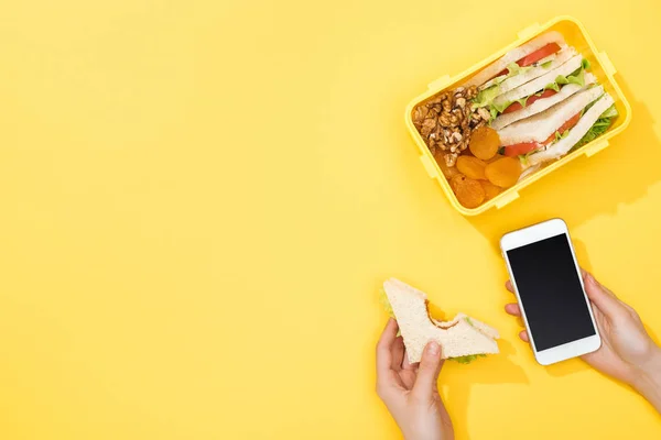 Vista recortada de la mujer sosteniendo sándwich y teléfono inteligente cerca de la lonchera con comida - foto de stock