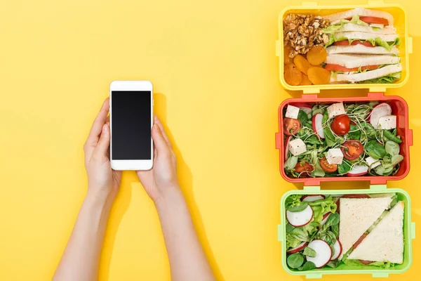 Vista recortada de la mujer sosteniendo teléfono inteligente cerca de loncheras con comida - foto de stock