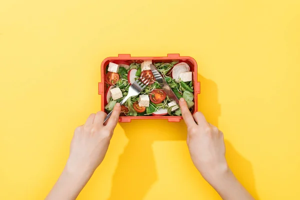 Обрезанный вид женщины, держащей вилку и нож, поедающей салат из коробки с обедом — стоковое фото