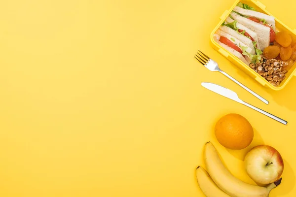 Top view of lunch box with sandwiches, nuts, dried apricots, near fork, knife, oranges and bananas — Stock Photo