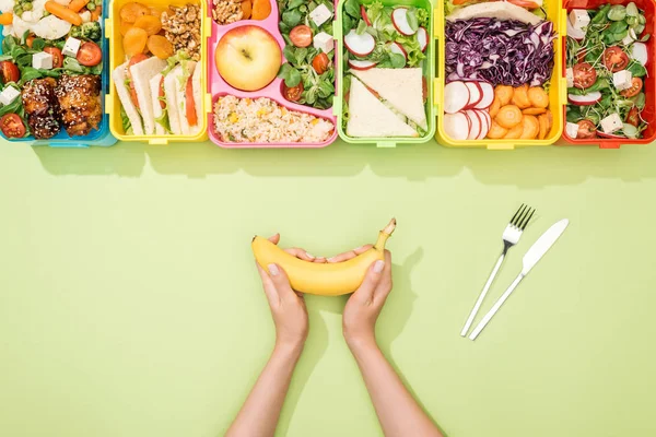 Cropped view of woman holding banana near fork, knife and lunch boxes with food — Stock Photo