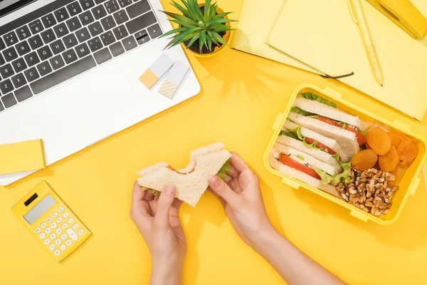 Cropped view of woman holding sandwich in hands near lunch box, laptop and office supplies — Stock Photo