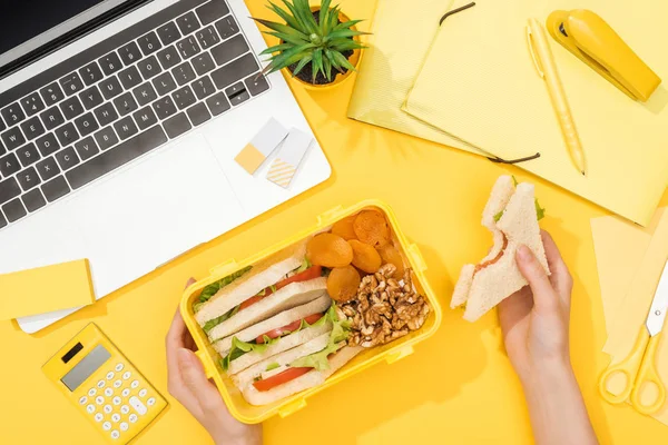 Обрезанный вид женщины, держащей сэндвич в руке рядом с обеденной коробкой, ноутбуком и офисными принадлежностями — стоковое фото