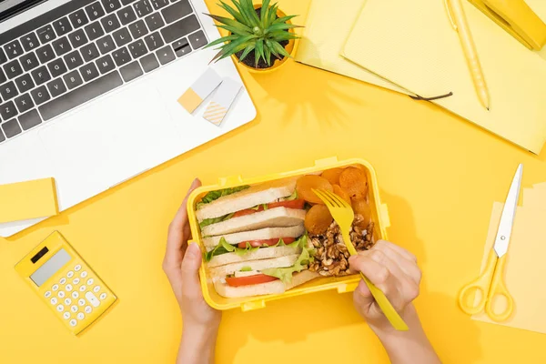 Cropped view of woman holding lunch box near laptop and office supplies — Stock Photo