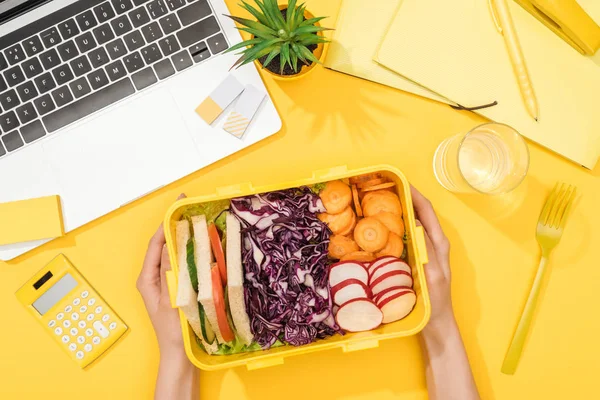 Cropped view of woman holding lunch box near laptop and office supplies — Stock Photo