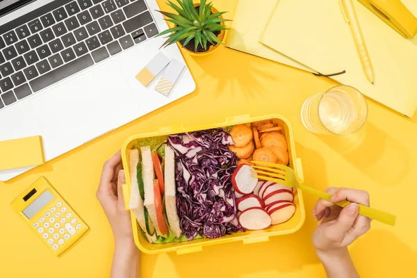 Cropped view of woman holding lunch box with food near laptop and office supplies — Stock Photo