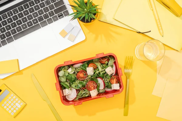 Вид на обеденную коробку с едой рядом с ноутбуком, стаканом воды и офисными принадлежностями — стоковое фото