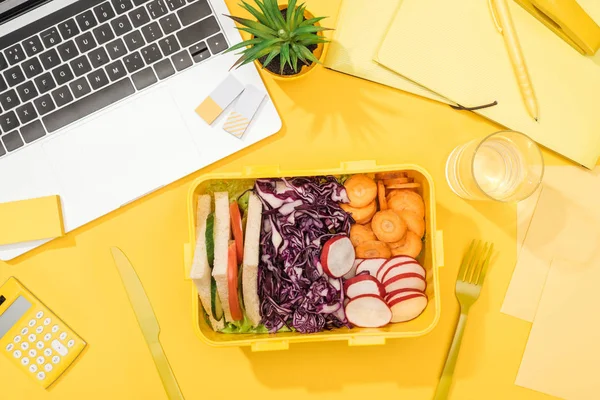 Вид на обеденную коробку с едой рядом с ноутбуком, стаканом воды и офисными принадлежностями — стоковое фото