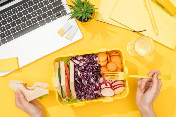 Vista recortada de la mujer sosteniendo sándwich en la mano cerca de la lonchera con comida, computadora portátil, vaso de agua y suministros de oficina - foto de stock