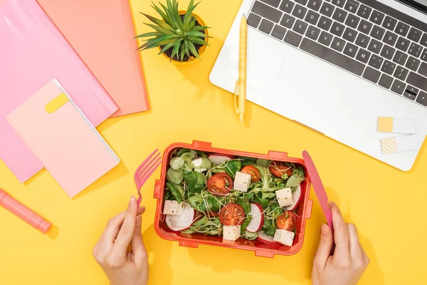 Cropped view of woman holding fork over lunch box with food near laptop and office supplies — Stock Photo
