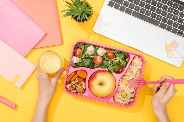 Cropped view of woman holding fork over lunch box with food near laptop and office supplies — Stock Photo