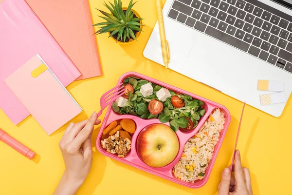 Cropped view of woman holding fork over lunch box with food near laptop and office supplies — Stock Photo